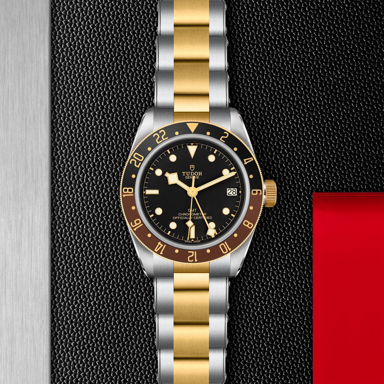 The TUDOR Black Bay GMT S&G wristwatch, featuring a gold and silver metal strap, is placed on a textured black surface. It showcases multiple time-zone features along with a black dial adorned with gold numerals and markers, a date window, and a rotatable bezel. On the right side, part of a small red card can be seen.