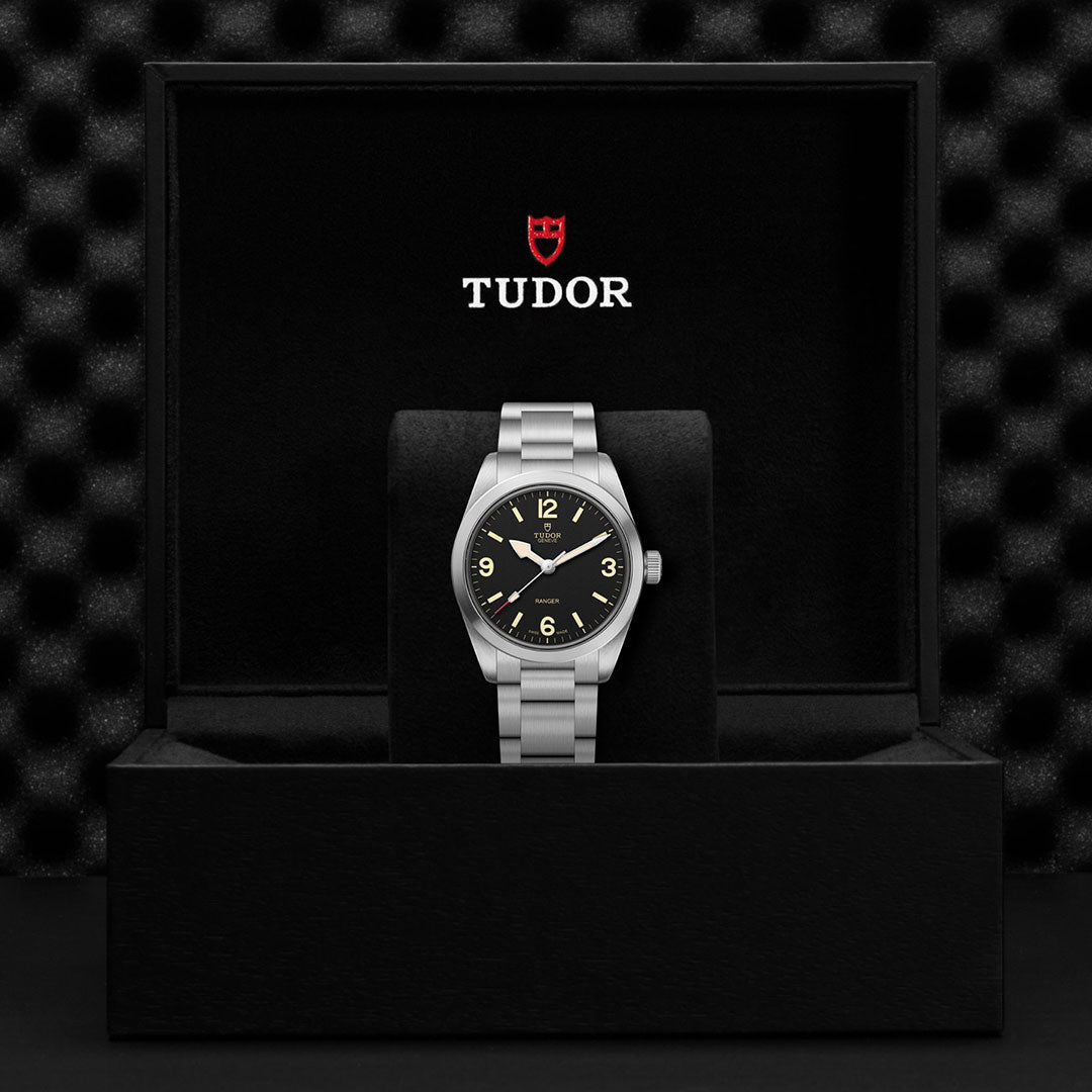 A TUDOR Ranger wristwatch with a black dial rests in an open black box displaying the TUDOR logo and name in white and red on the inside lid, set against a textured dark background.