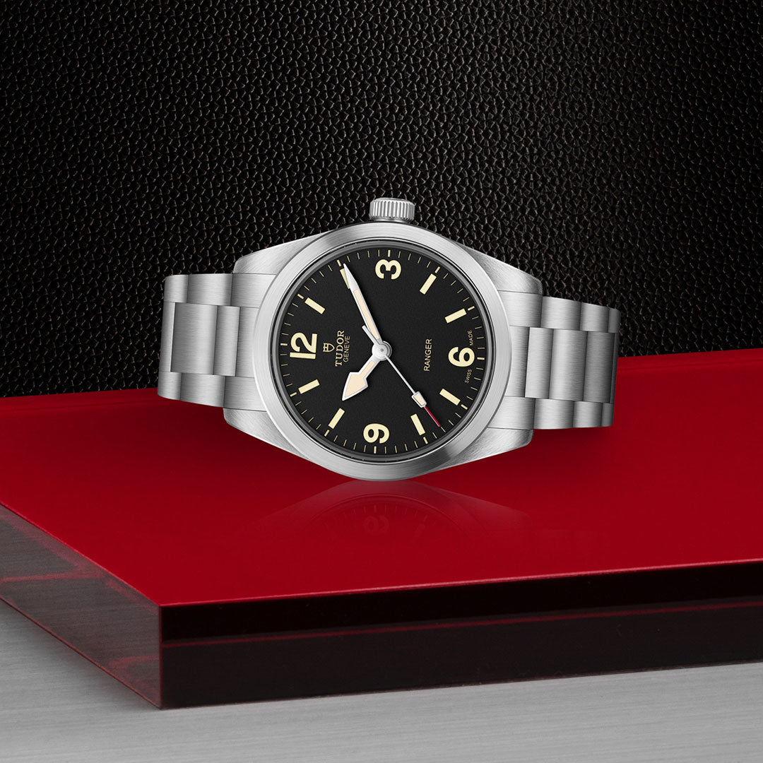 A TUDOR Ranger watch by TUDOR, featuring a silver stainless steel band and black dial, shows 10:10 atop a glossy red surface with a textured black background, powered by the Manufacture Calibre MT5400 movement.