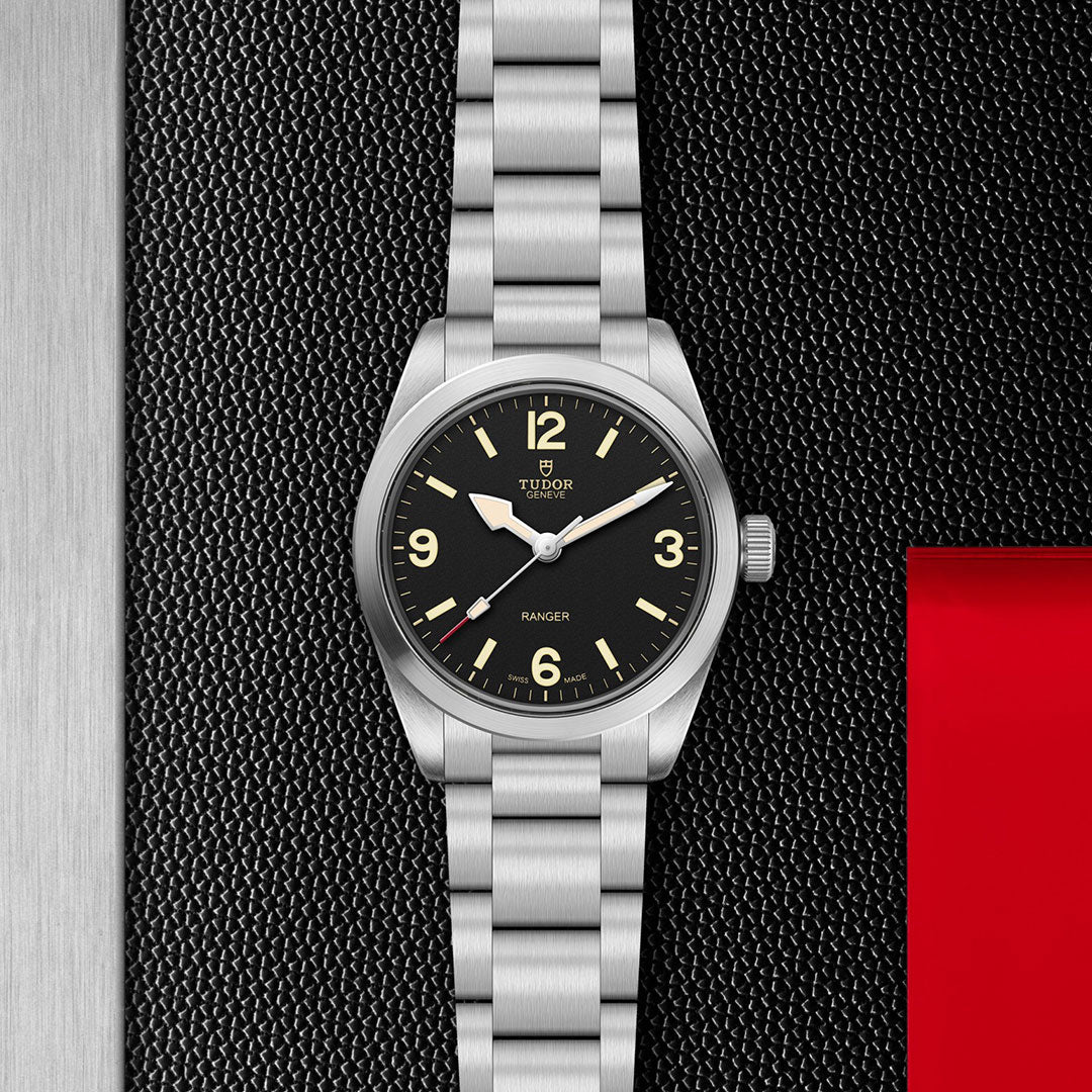 The TUDOR Ranger by TUDOR is a stainless steel expedition watch with a black dial, luminous hands and hour markers, powered by the Manufacture Calibre MT5400, shown on a textured black surface beside a red accent.