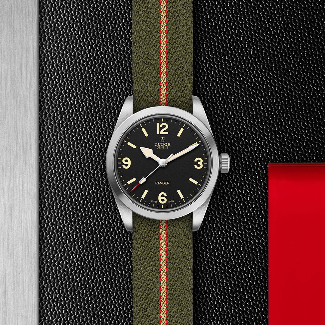 The TUDOR Ranger by TUDOR features a black dial, beige numerals, and an olive green fabric strap with a red stripe. Set against a textured black and silver background with red accents, it embodies the spirit of modern exploration with Manufacture Calibre MT5400.