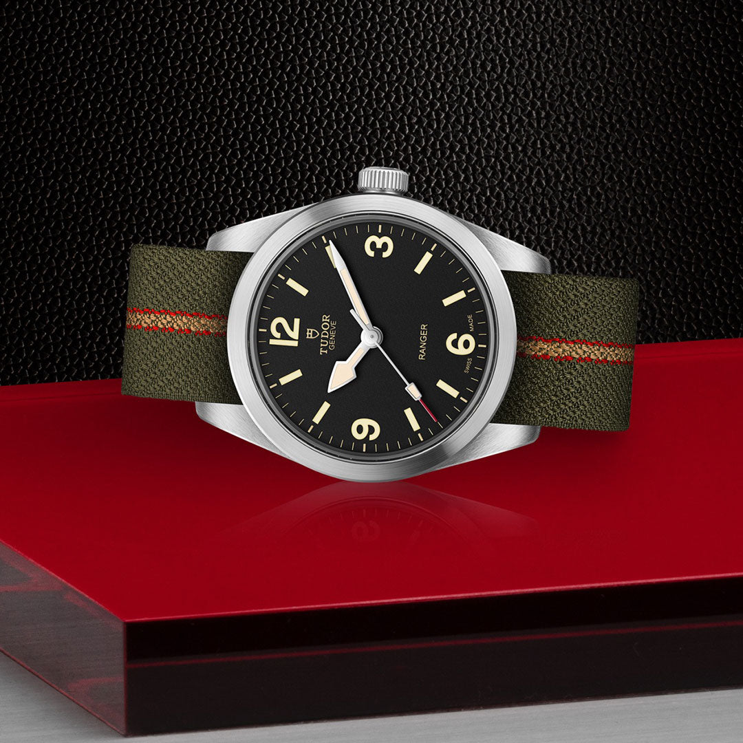 The TUDOR Ranger by TUDOR features a black dial, luminous markers, an olive green strap with a red stripe, and runs on the Manufacture Calibre MT5400—shown here on a striking red and black textured surface.