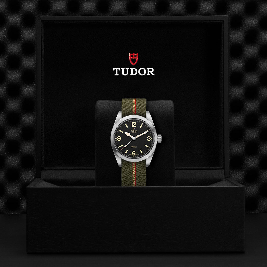 The TUDOR Ranger, featuring a green fabric strap and black dial, is showcased in an open black TUDOR box with the brand logo on the inside lid. A textured dark background emphasizes this modern TUDOR expedition watch.