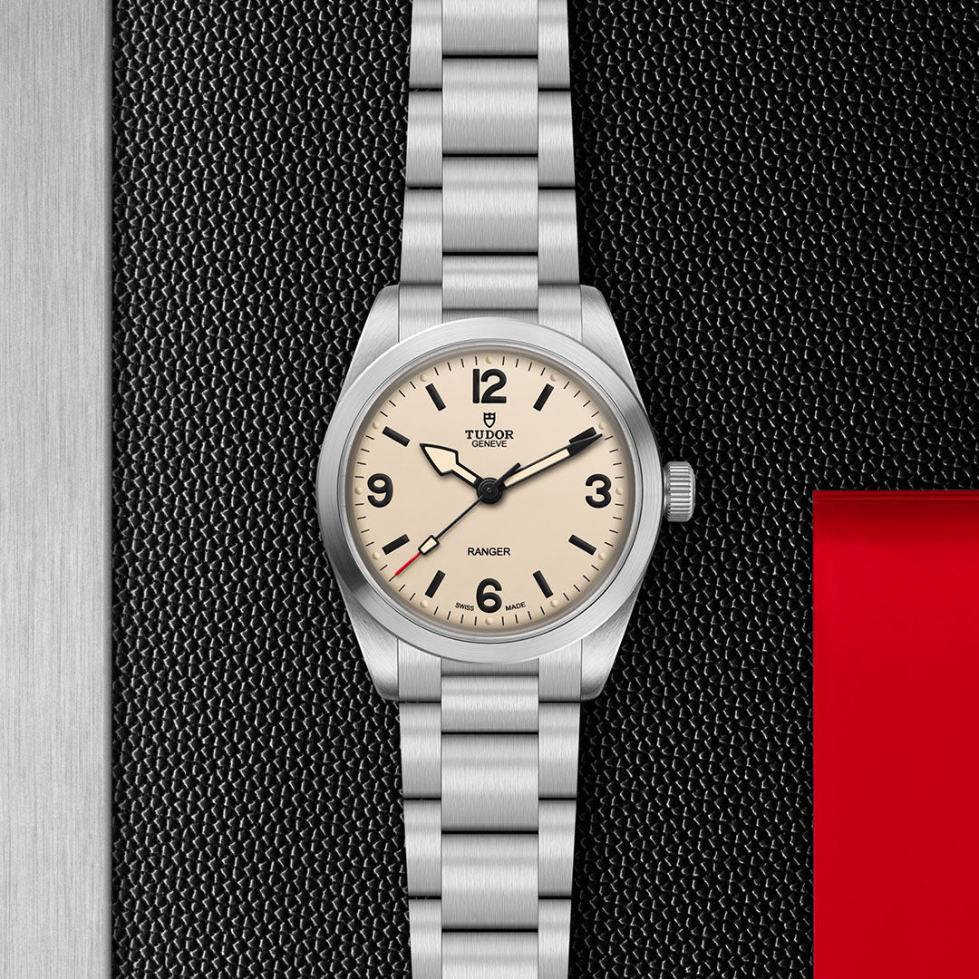 The TUDOR Ranger by TUDOR features a beige dial, black hour markers, metal bracelet, and is powered by Manufacture Calibre MT5400. Displayed on a textured black surface with a red accent on the right.