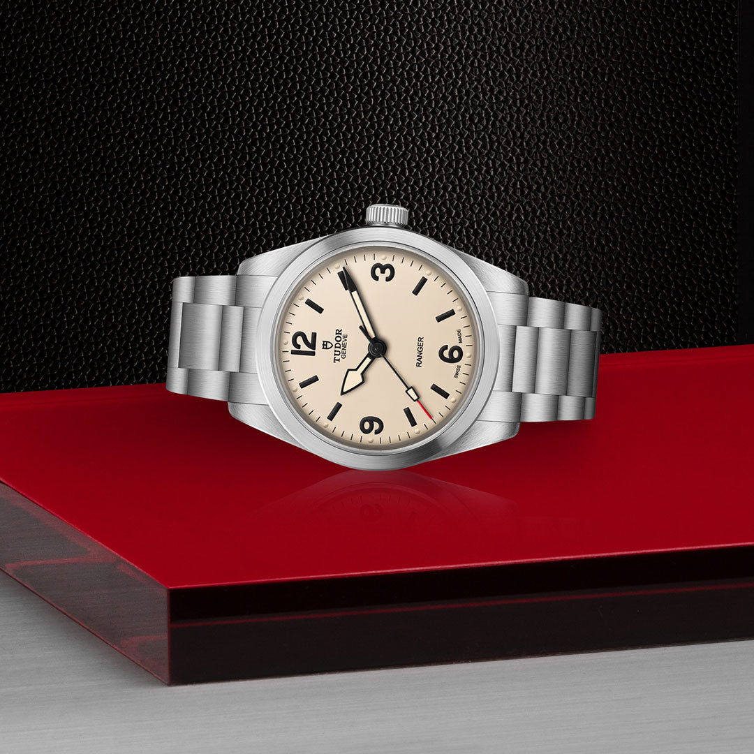 A TUDOR Ranger watch with a cream dial, black hour markers, and a metal bracelet is displayed on a glossy red surface against a textured black background.