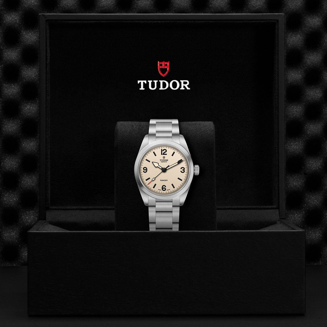 A TUDOR Ranger wristwatch with a silver metal band and beige dial is displayed upright in an open black box, featuring the red TUDOR logo and name on the inside lid.
