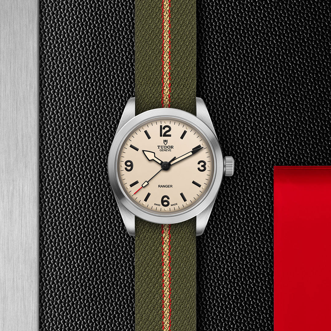 The TUDOR Ranger by TUDOR features a cream dial, black numerals, and a green fabric strap with a red stripe. Shown on a textured black and metal background with a red accent, it’s powered by the Manufacture Calibre MT5400.