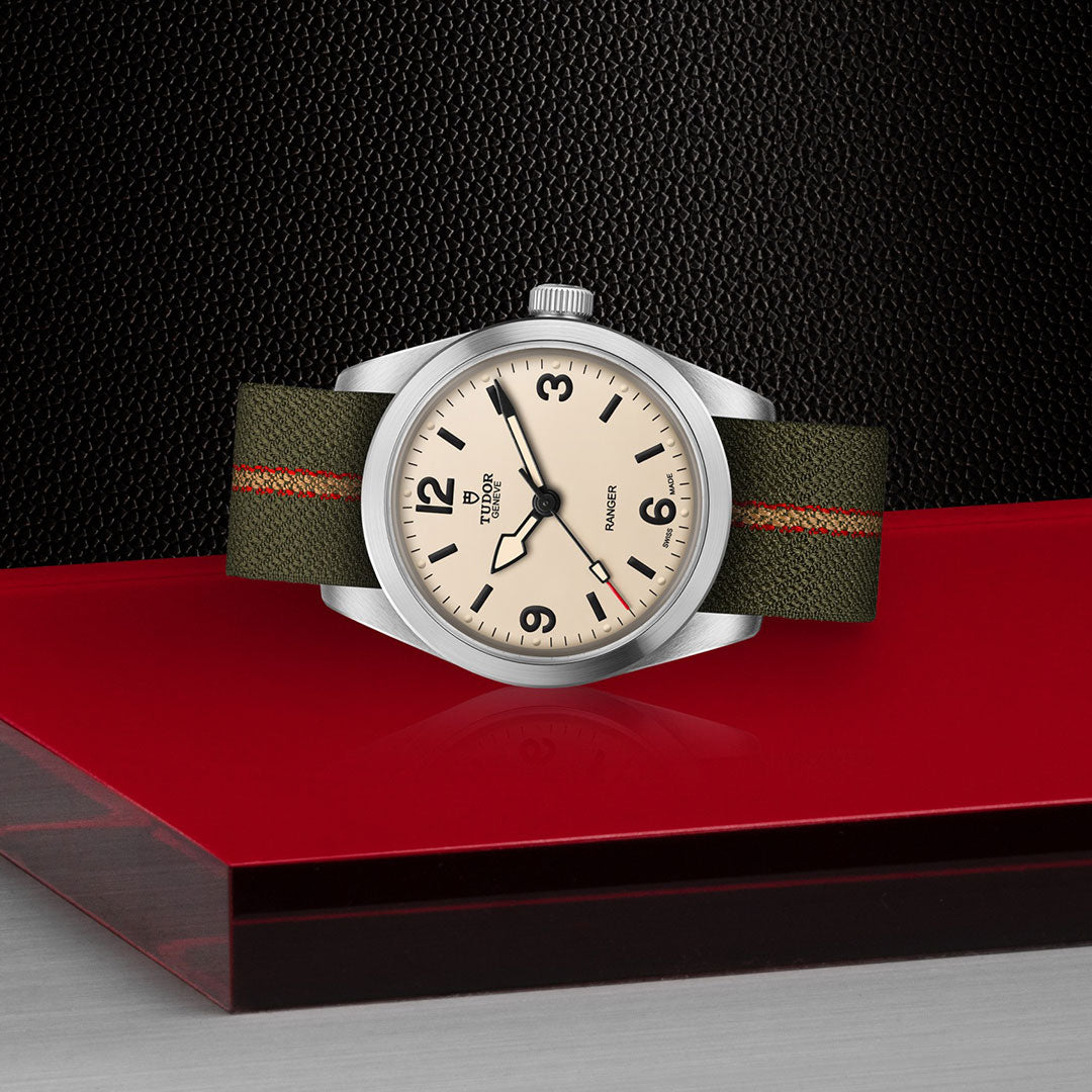 The TUDOR Ranger by TUDOR features a beige dial, black numerals, and a green fabric strap with a red stripe, shown on glossy red and gray with a textured black background. It is powered by the Manufacture Calibre MT5400.