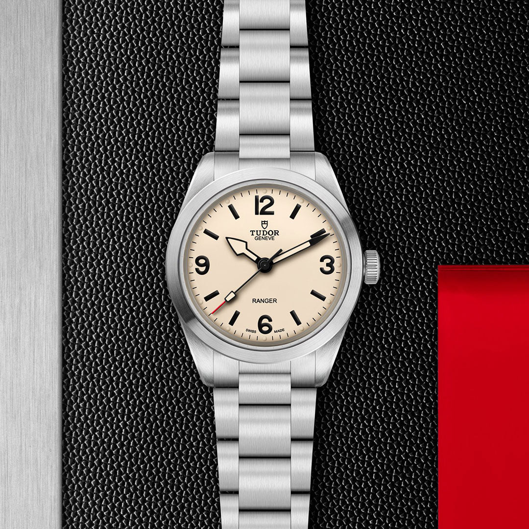 The TUDOR Ranger by TUDOR features a stainless steel bracelet, beige dial with black numerals, a red-tipped second hand, and Manufacture Calibre MT5400. Shown on a textured black background with red accent and silver edge.