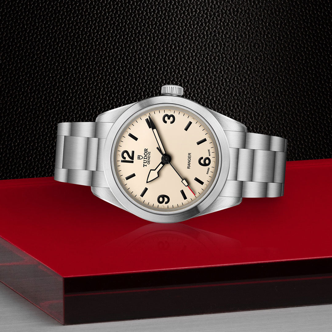 The TUDOR Ranger by TUDOR, inspired by the original expedition watch, features a silver case with a cream dial and black numerals, showcased on a glossy red and clear platform with a textured black background.