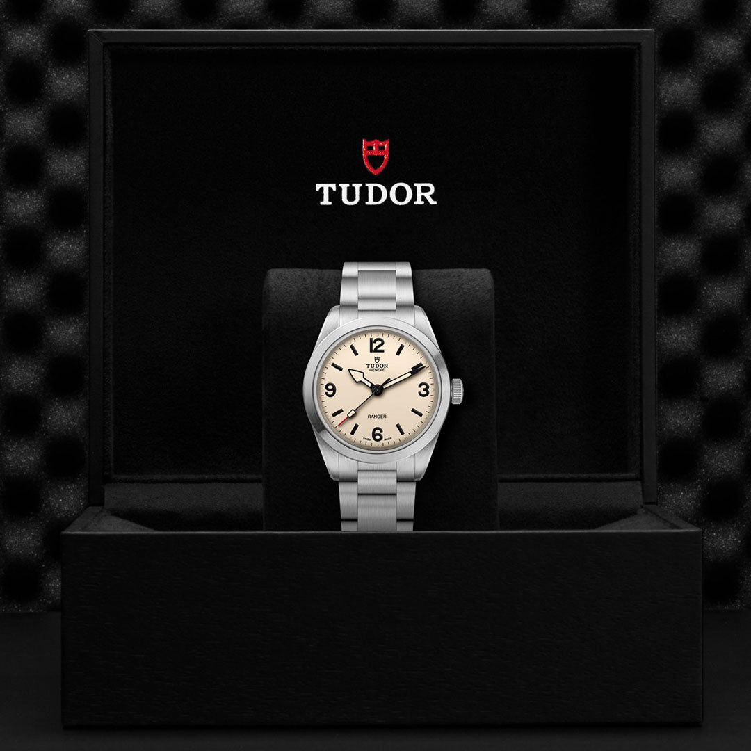 A TUDOR Ranger wristwatch with a silver case, beige dial, and black numerals is displayed in an open black box featuring the TUDOR logo and shield inside the lid—a classic expedition watch from TUDOR designed for adventure.