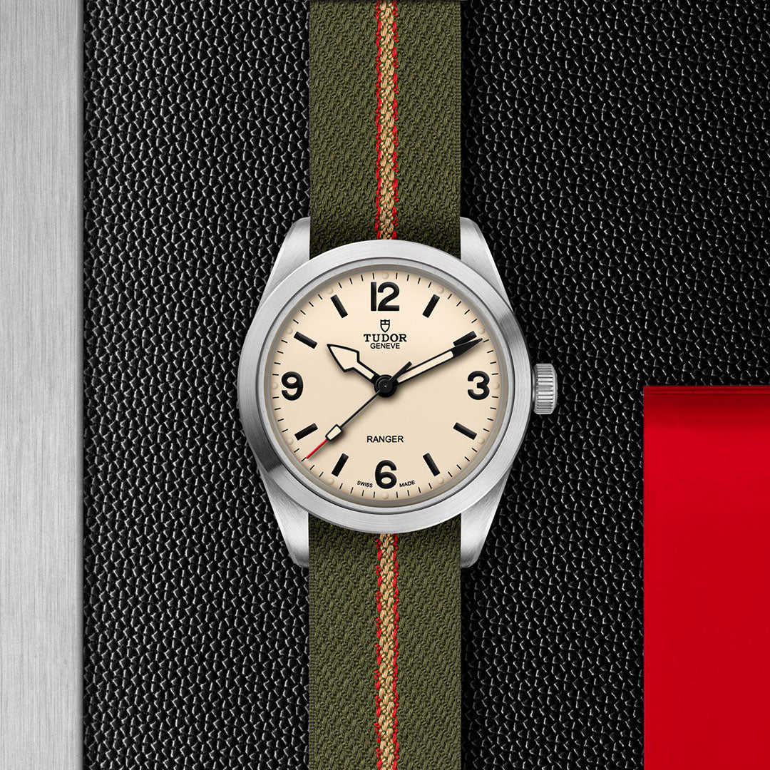 The TUDOR Ranger expedition watch features a beige dial with black numerals and hands, paired with an olive green fabric strap with a red stripe, and is displayed on a textured black surface with silver and red accents. Brand: TUDOR.