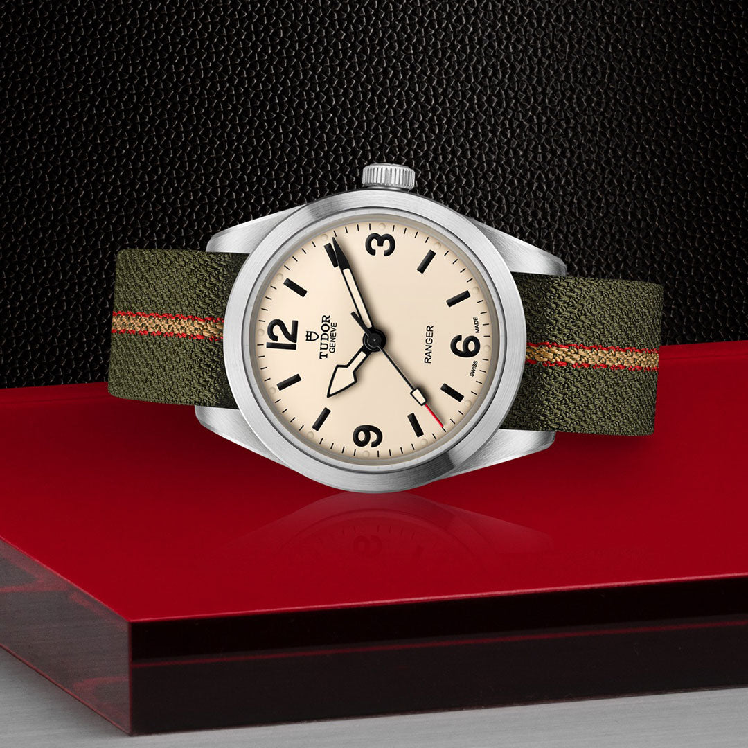 The TUDOR Ranger by TUDOR features a cream dial with black numerals and an olive green fabric strap with red and beige stripes, displayed on a glossy red rectangle against a textured black background.