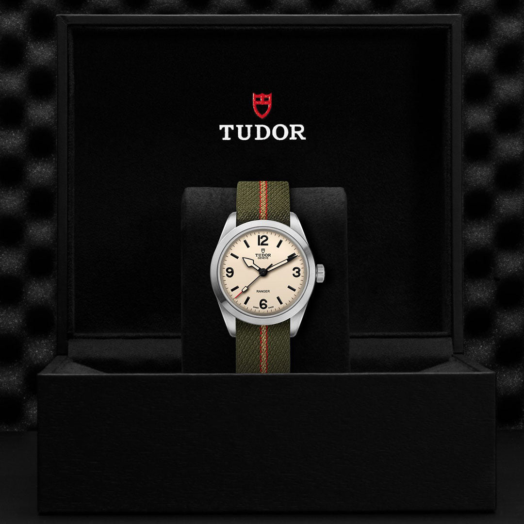 The TUDOR Ranger by TUDOR, featuring a beige dial and green fabric strap, is displayed upright in a black presentation box with the Tudor logo inside the lid.