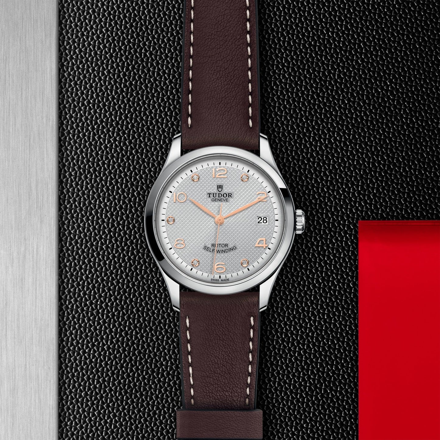 The TUDOR 1926 watch, a creation by the renowned TUDOR brand, features a silver casing and a distinctive white textured dial adorned with orange hour markers and a date display at the 3 o'clock position. Its elegant brown leather strap with white stitching provides an appealing contrast against the textured black backdrop accentuated by a touch of red. This timepiece is perfect for aficionados of mechanical watches.