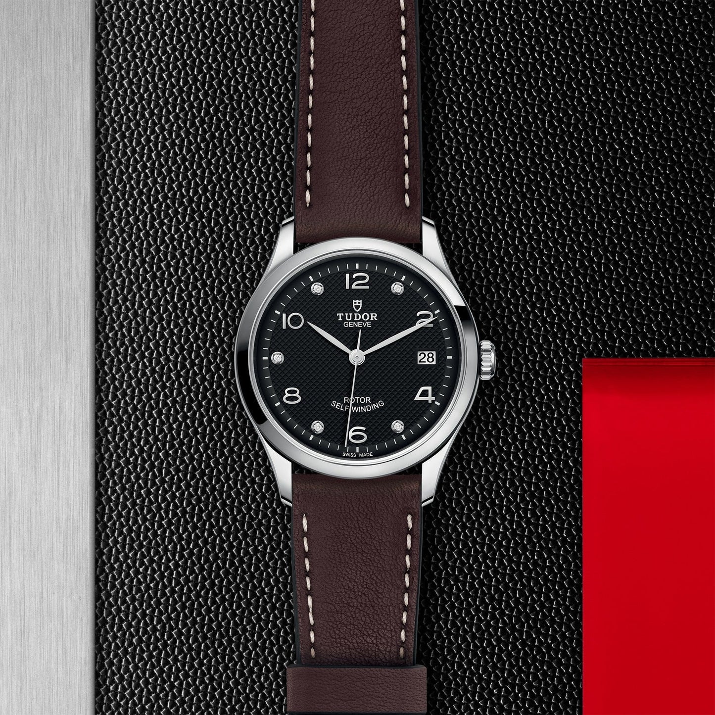 The TUDOR 1926 wristwatch is displayed against a textured dark backdrop, highlighting its stainless steel edge and red accent. This luxurious mechanical watch features a black dial, complemented by a brown leather strap and silver casing. It includes Arabic numerals and a date function.