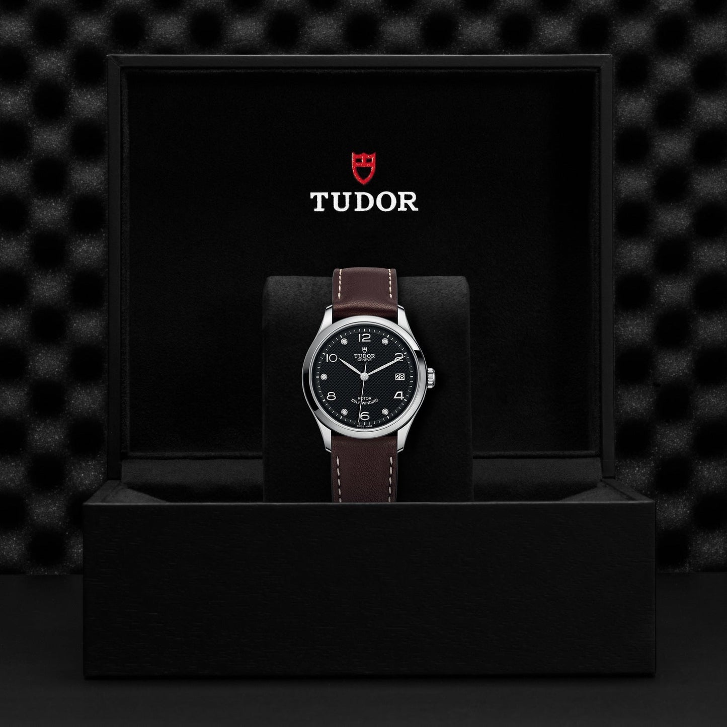 A TUDOR 1926 wristwatch featuring a diamond-set black dial and brown leather strap is elegantly displayed upright in a sleek black presentation box. The TUDOR logo enhances the open box lid, beautifully complementing the textured black background that frames this exquisite mechanical timepiece.