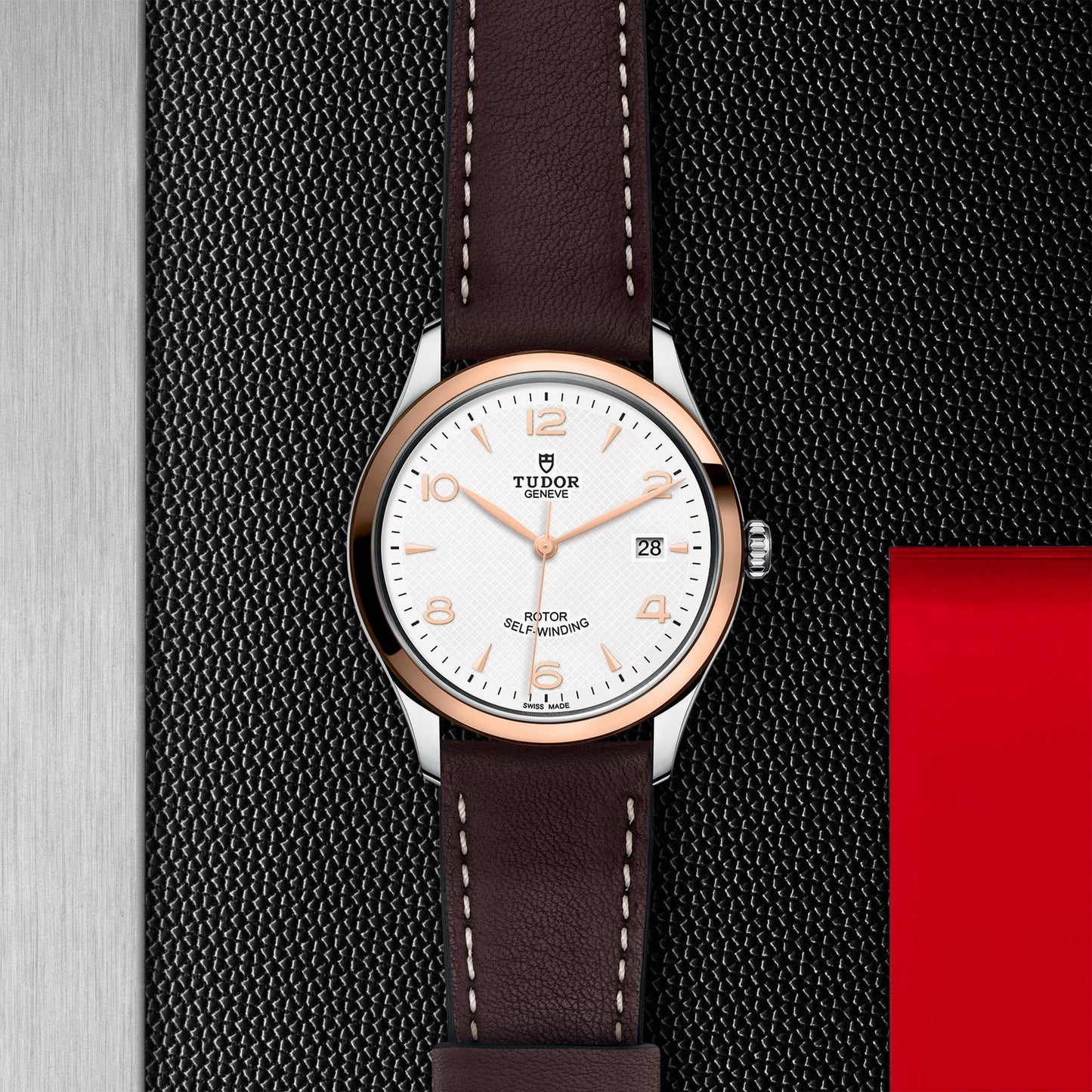 The TUDOR 1926 wristwatch from TUDOR features a brown leather strap and a white dial adorned with gold hour markers and hands. This sophisticated mechanical timepiece includes a date display at the 3 o'clock position, showcased against a textured black surface with an eye-catching metallic and red background.