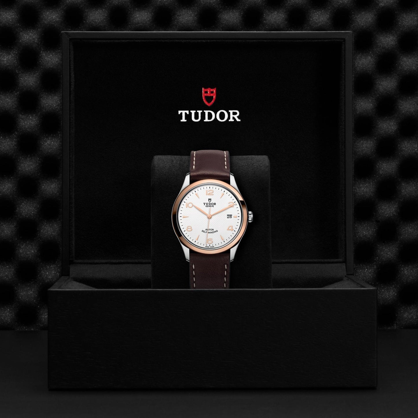 A TUDOR 1926 wristwatch, adorned with a white dial and brown leather strap, is showcased in an open black box. The TUDOR logo and name are elegantly presented inside the lid against a textured black background.