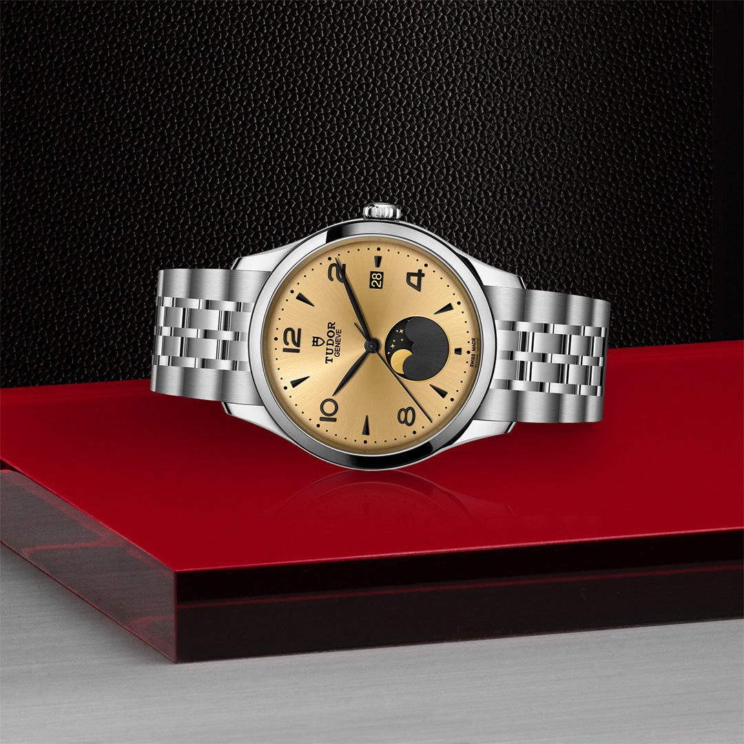 The TUDOR 1926 Luna wristwatch, featuring a gold dial, moonphase display, and silver metal bracelet, is showcased on a glossy red and black surface against a textured black background—a symbol of classic elegance.
