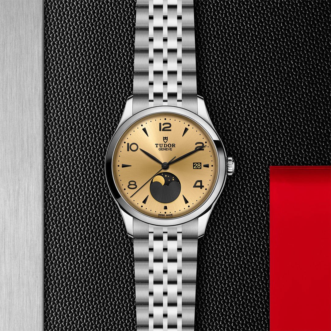 The TUDOR 1926 Luna by TUDOR features a gold dial, moonphase, date window, and silver bracelet—set against a textured black and metallic background with a red accent—ideal for mechanical watch enthusiasts.