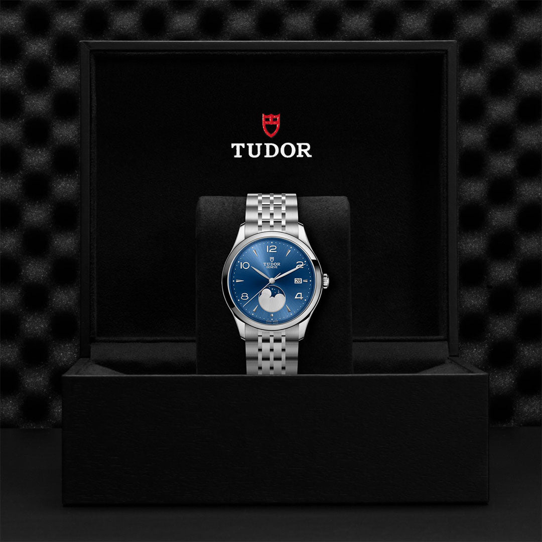 The TUDOR 1926 Luna, a silver wristwatch with a blue dial, is showcased upright in a black TUDOR box featuring a textured interior and the brand’s logo on the inner lid.