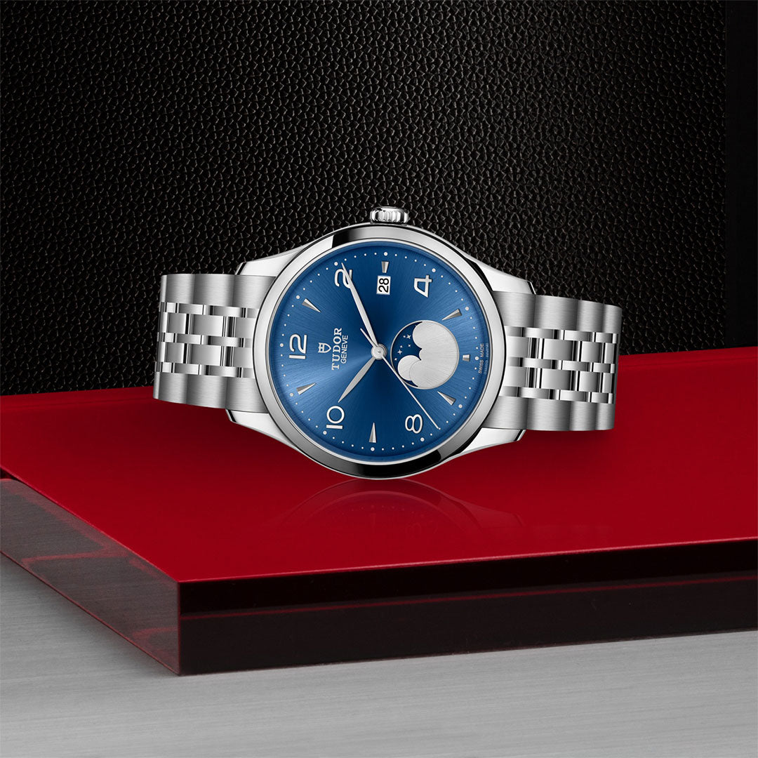 The TUDOR 1926 Luna, a silver wristwatch with a blue dial and moonphase display, rests on a glossy red and black surface against a textured black background.