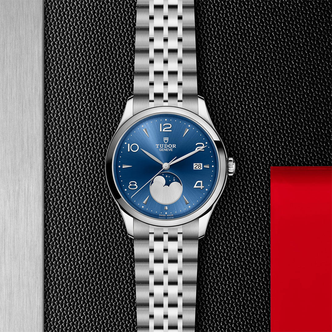 The TUDOR 1926 Luna wristwatch features a striking blue dial, moon phase display, and metal link bracelet, elegantly displayed on a textured black and red background—a timeless piece for classic watch enthusiasts.
