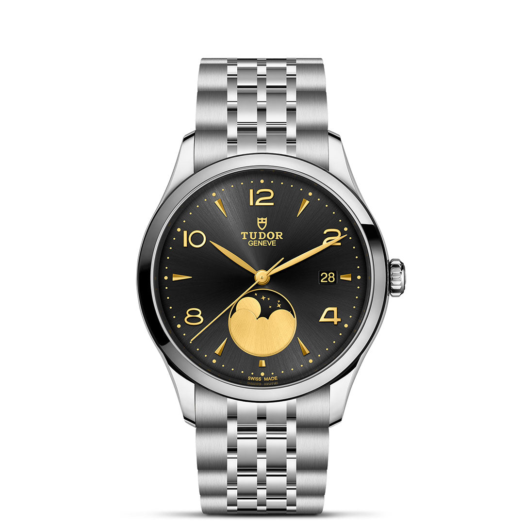 The TUDOR 1926 Luna by TUDOR is a silver wristwatch with a black and gold dial, gold hands and numerals, moon phase at 6 o’clock, date window at 3 o’clock, and a silver metal bracelet strap.