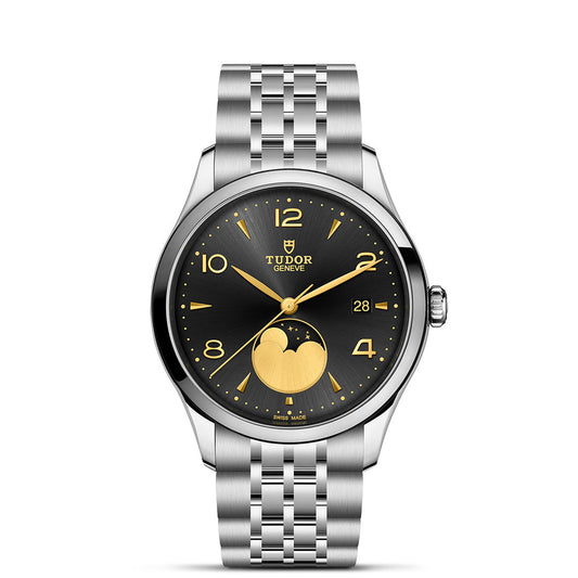 The TUDOR 1926 Luna by TUDOR is a silver wristwatch with a black and gold dial, gold hands and numerals, moon phase at 6 o’clock, date window at 3 o’clock, and a silver metal bracelet strap.