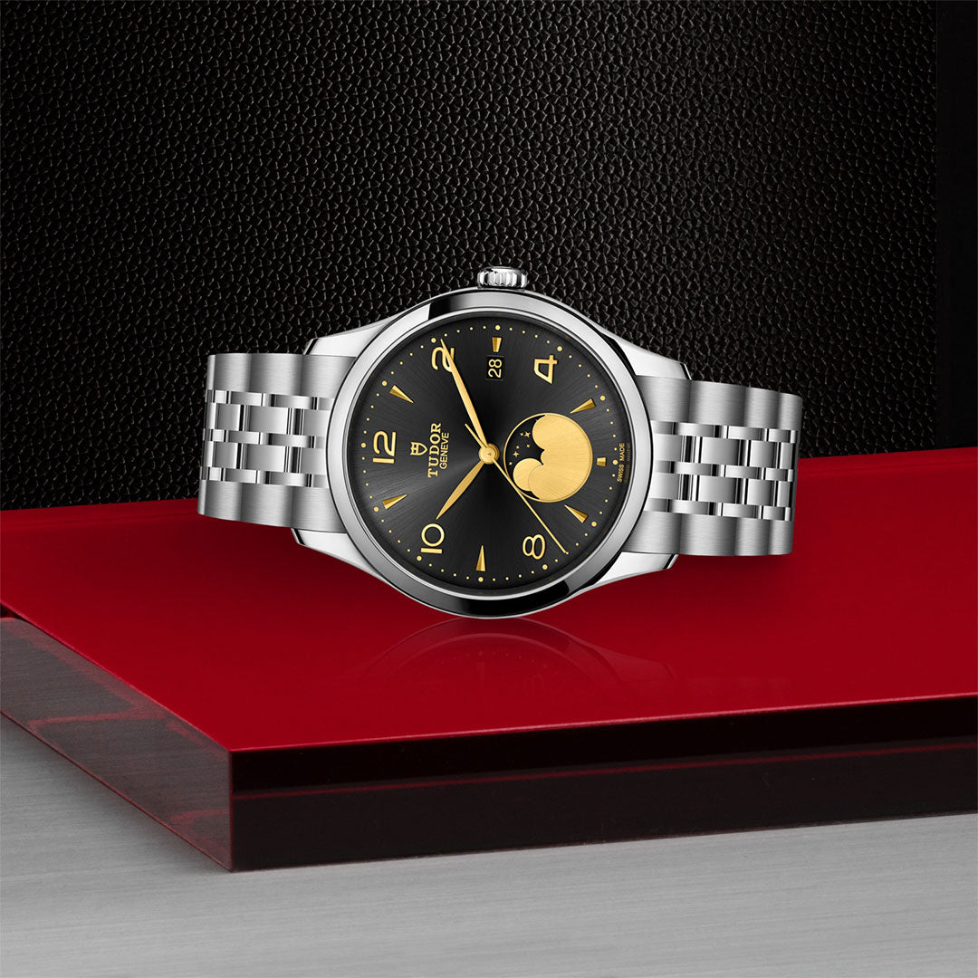 The TUDOR 1926 Luna wristwatch features a black and gold moon phase dial, mechanical movement, and stainless steel bracelet, displayed on a glossy red and black surface with a textured black backdrop.