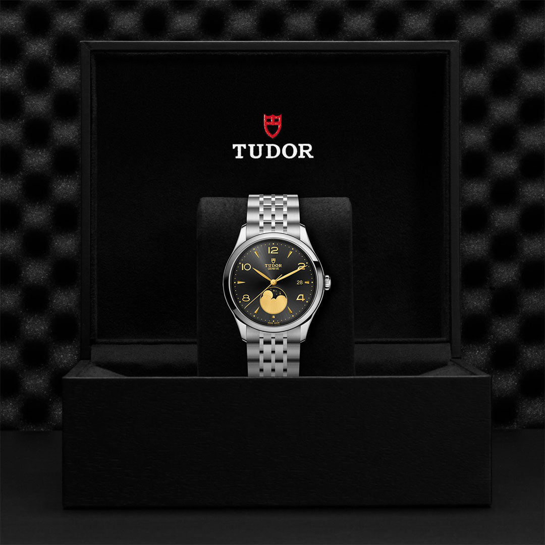A TUDOR 1926 Luna wristwatch with a black dial and metal bracelet is displayed upright in an open black box featuring the TUDOR logo, standing out elegantly against a textured, patterned backdrop.