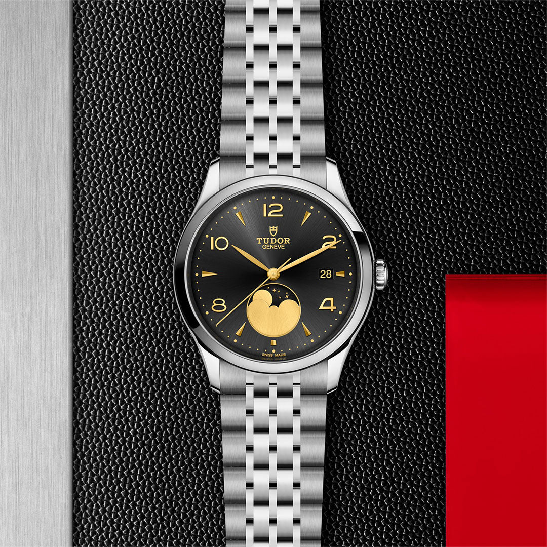 The TUDOR 1926 Luna wristwatch by TUDOR, with a black and gold dial, moonphase display, date window, and stainless steel bracelet, is shown on a textured black and metallic background with a red accent for timeless elegance.