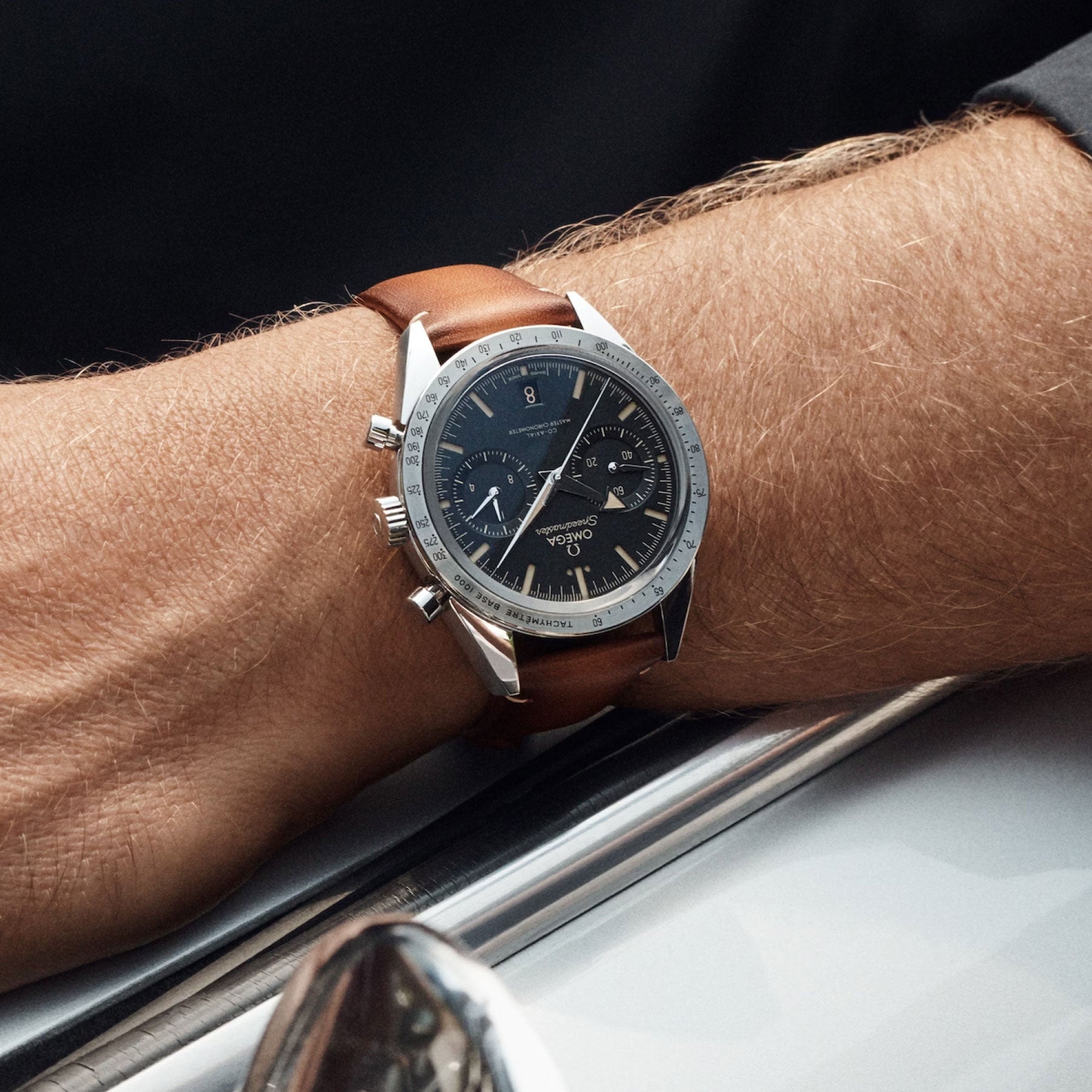 Close-up of a wrist showcasing an OMEGA Speedmaster 57 Co-Axial Master Chronometer Chronograph 40.5mm watch, featuring a classic stainless steel look, complemented by a brown leather strap and a dark dial with two subdials. The chronograph is positioned on a person's arm resting against what appears to be the metallic edge of a car window.