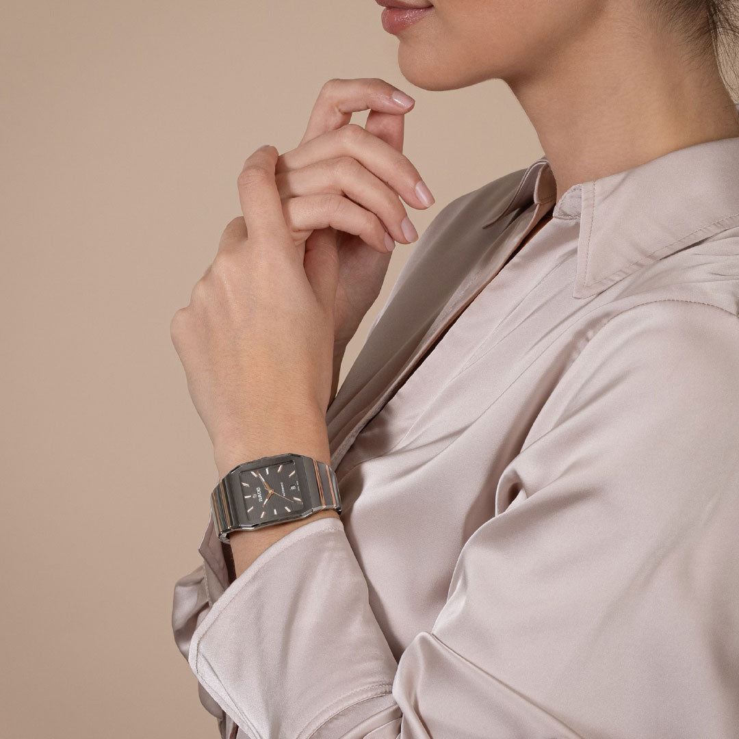 A woman in a light beige satin blouse is shown from the shoulders up, hands gently clasped, wearing the RADO Anatom Automatic 32.5mm watch with sapphire crystal on her left wrist against a neutral background.