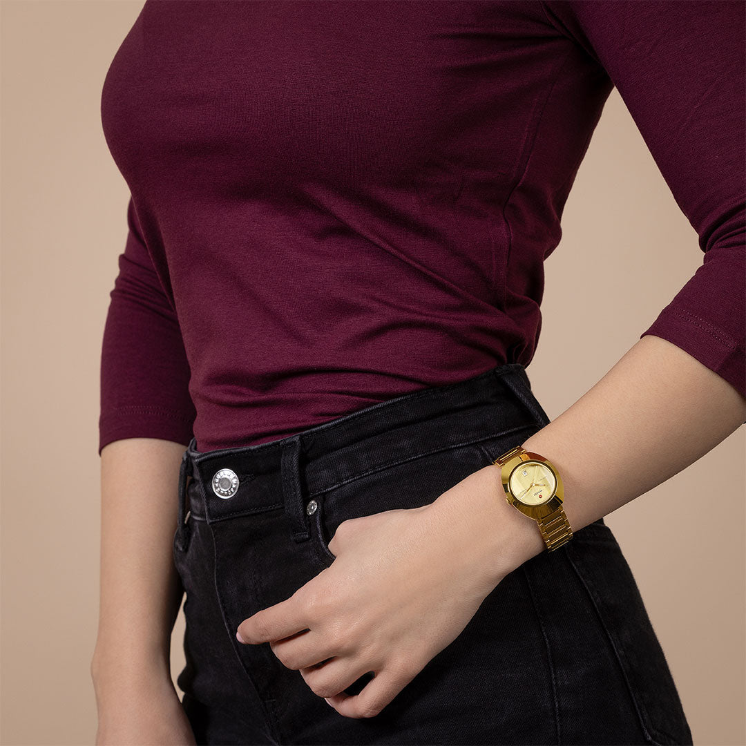 A person in a maroon long-sleeve top and black jeans, left hand in pocket, displays the RADO Diastar Original Automatic 30mm Watch by RADO on their wrist against a beige background.