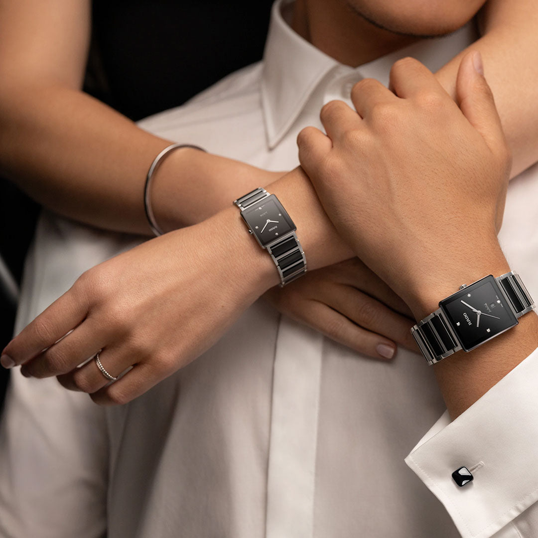 Two people pose with arms crossed, each wearing a matching RADO Integral Diamonds Quartz 23mm watch. The rectangular black-faced watches by RADO feature sapphire crystal displays; one wearer sports a white shirt and cufflinks, the other a thin bracelet and ring.