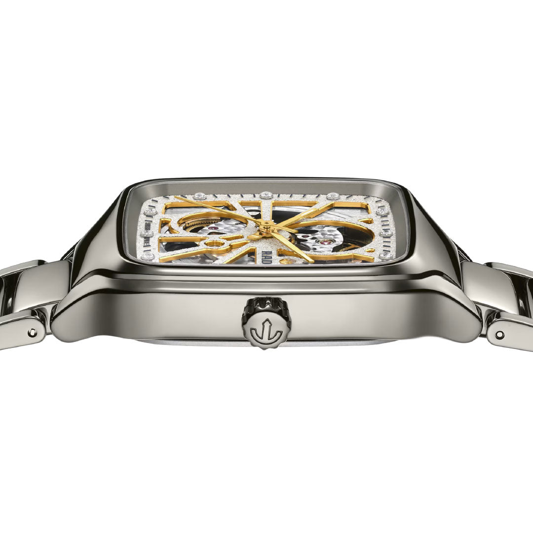 Side view of the RADO True Square Automatic Open Heart Sparkling 38mm watch, featuring a polished metal case, skeleton dial with gold accents, metal bracelet, and a crown engraved with the RADO logo.