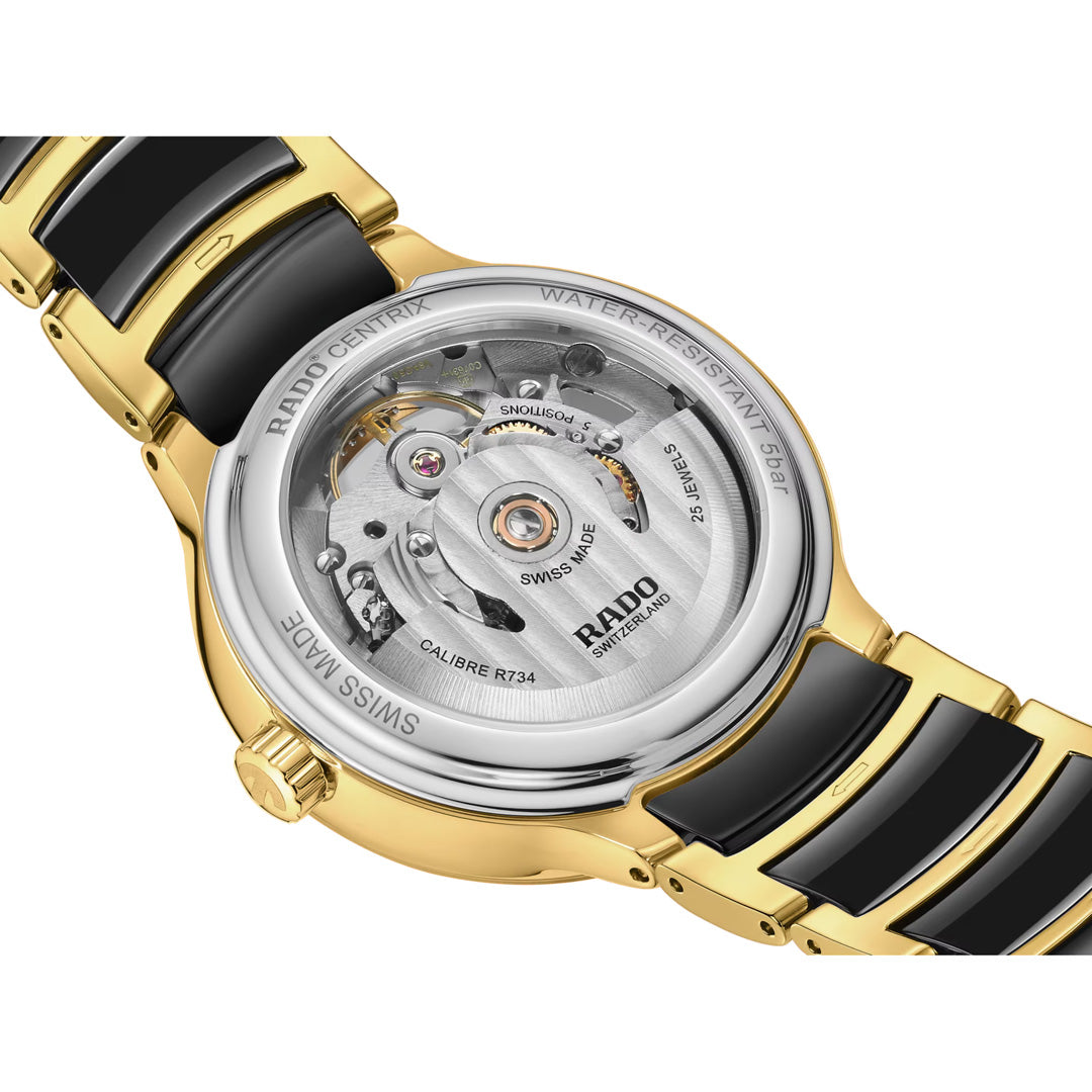 The image features the back of the RADO Centrix Open Heart Automatic 35mm Watch, highlighting its automatic movement through a transparent case back. The gold and black design reveals intricate mechanisms along with engraved details about water resistance and model information, enhancing the refined elegance characteristic of RADO timepieces.