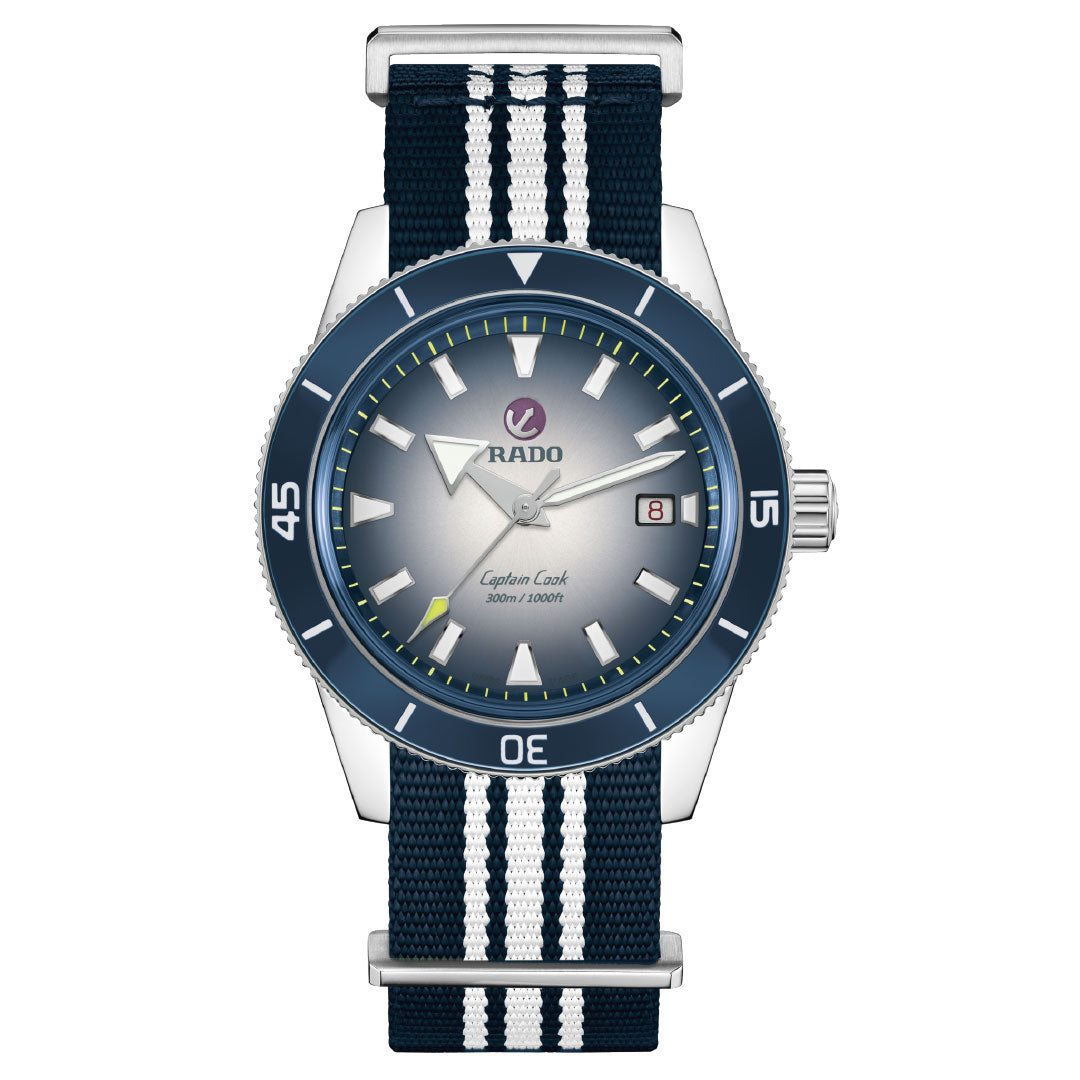 The RADO Captain Cook Automatic 42mm Watch features a stainless steel case, blue and white fabric strap, blue bezel, gradient gray dial with luminous markers, 80-hour power reserve, date at 3 o'clock, and "Captain Cook" above 6 o'clock.