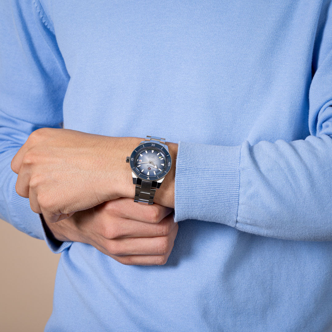 A person in a light blue long-sleeve shirt wears a RADO Captain Cook Automatic 42mm Watch on their left wrist, holding it with their right hand. The image shows the torso and hands, not the face.