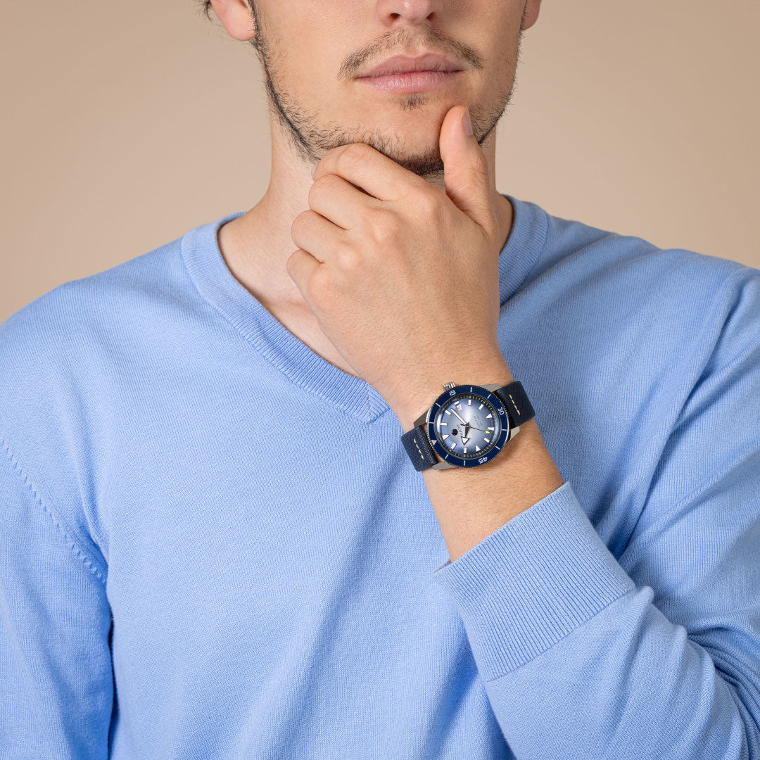 A man in a light blue sweater thoughtfully touches his chin, wearing the RADO Captain Cook Automatic 42mm Watch on his left wrist. The background is plain beige.