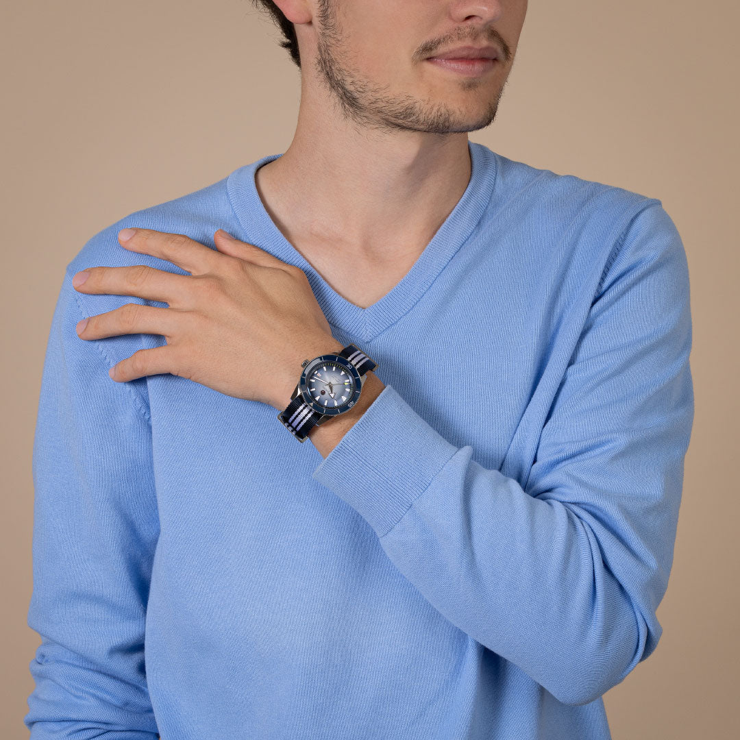 A man in a light blue long-sleeve shirt poses with one hand on his shoulder, showcasing the RADO Captain Cook Automatic 42mm Watch with a striped band against a plain beige background.