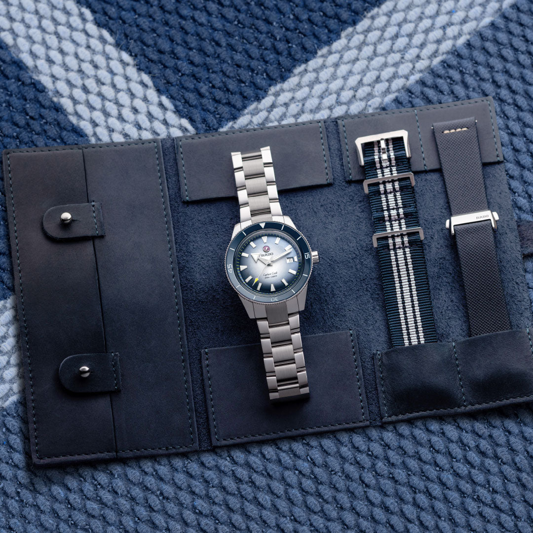 The RADO Captain Cook Automatic 42mm Watch, offering an 80-hour power reserve, is presented in a dark leather case with two interchangeable straps—striped fabric and textured rubber—on a blue patterned surface.