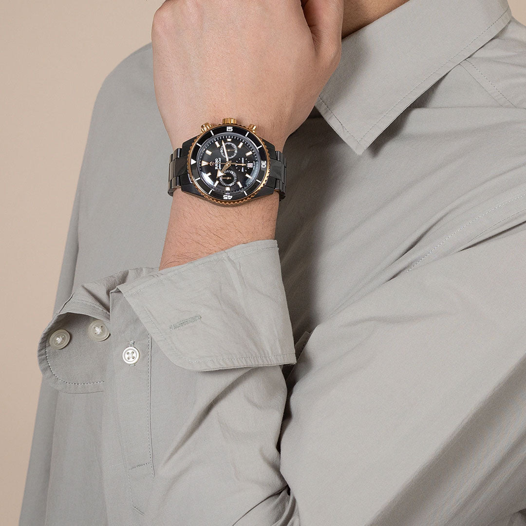 A person in a light gray, long-sleeved shirt holds their hand to their face, showcasing the RADO Captain Cook High-Tech Ceramic Automatic Chronograph 43mm Watch on their wrist.
