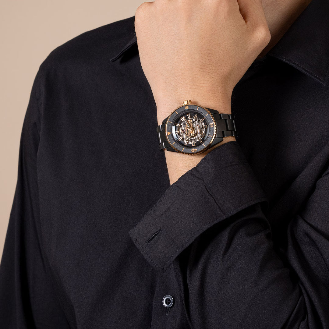 A person in a black dress shirt poses with their hand near their face, showcasing the RADO Captain Cook High-Tech Ceramic Skeleton Automatic 43mm Watch in bold black and gold.