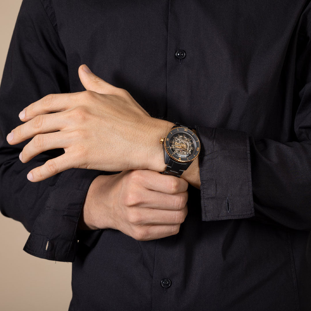 A person in a black dress shirt adjusts their cuff, displaying the RADO Captin Cook High-Tech Ceramic Skeleton Automatic 43mm Watch with a bold black and gold design on their left wrist.