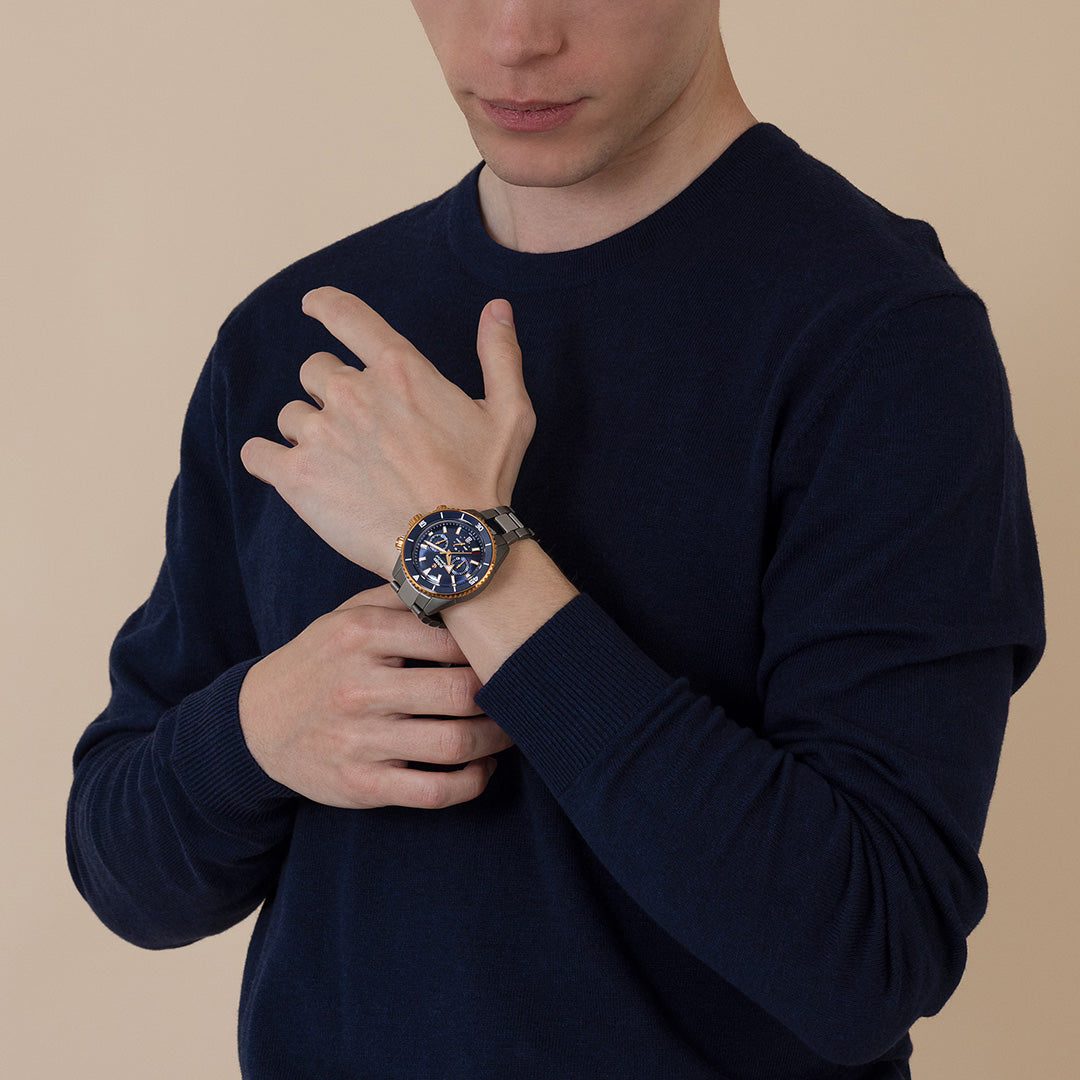 Wearing a navy sweater, a person adjusts the RADO Captain Cook Hi-Tech Ceramic Chronograph Automatic 43mm Watch with a striking blue face and gold bezel on their left wrist, standing against a plain beige background.