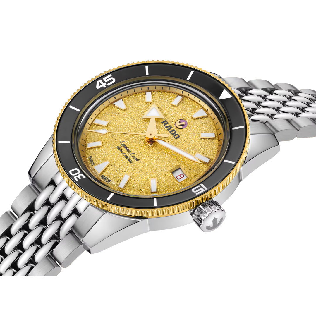 A detailed close-up of the RADO Captain Cook Automatic 39mm Watch highlights its gold textured dial, silver-toned hour markers, black and gold bezel, date window, and elegant silver metal link bracelet. The crown showcases a small anchor emblem, a signature detail from the iconic RADO Captain Cook collection.