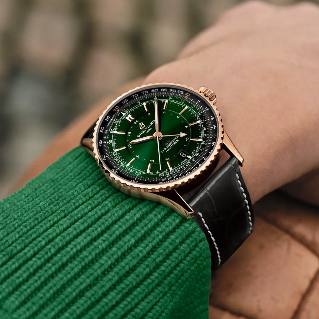 A wrist wearing a BREITLING Navitimer Automatic GMT 41 by Breitling, featuring a green dial, black leather strap, and gold bezel, styled with a green sweater against a softly blurred outdoor background.